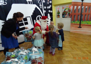Święty Mikołaj wręcza dzieciom prezenty. Nauczycielka podaje torebkę z prezentem.